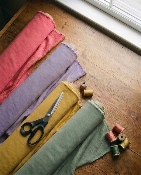 Flat lay materiałów pracowni SELVARRA — rolki dresówki w paletach koral, lawenda, żółć, sage z nożyczkami i szpulami nici